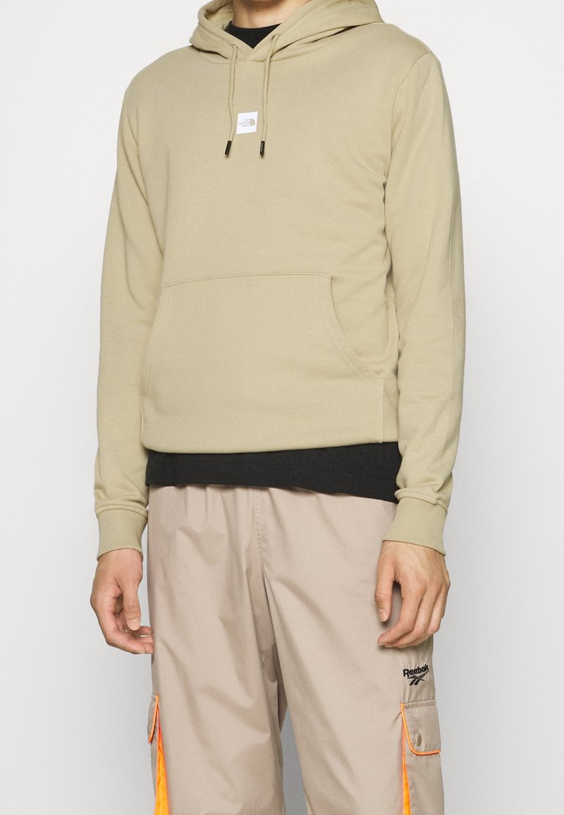 Felpa beige con cordini regolabili, tasca frontale e toppa con logo. Indossata sopra una maglietta nera, abbinata a pantaloni chiari con dettagli arancioni.