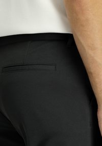 Pantaloni neri in tessuto testurizzato, caratterizzati da una vestibilità slim, una tasca posteriore discreta e un elastico in vita, parzialmente visibile all'altezza dell'anca.