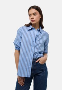 Chemise rayée bleue avec un col boutonné, présentant des rayures verticales bleues et blanches, des manches retroussées et une coupe décontractée.