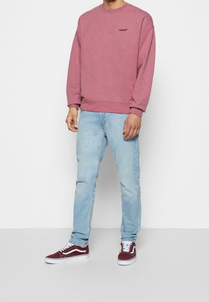 Felpa rosa con un logo nero, abbinata a jeans azzurri chiari e sneakers di camoscio rosse con dettagli e lacci bianchi.