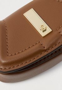 Colț de accesoriu din piele maro cu cusături detaliate și o placă metalică aurie rectangulară, cu un logo dublu „B” ridicat.