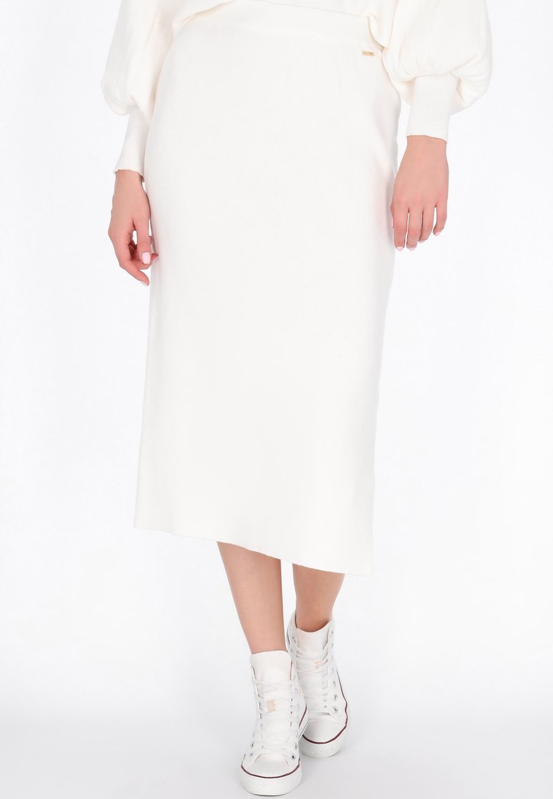 Biała dzianinowa spódnica midi o prostym kroju z delikatnym asymetrycznym dołem, w połączeniu z białymi sneakersami za kostkę. Gładka struktura, minimalistyczny design.