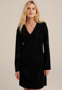 Black velvet dress with a V-neckline, long sleeves, and a relaxed fit. The fabric has a subtle sheen and a textured surface.