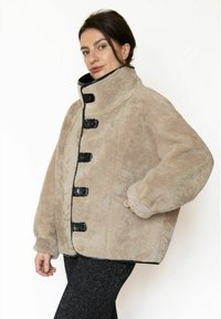 Beige jakke i imiteret pels med høj krave, sorte knapper og kontrastærmer. Har en afslappet, oversize pasform med brede ærmer.