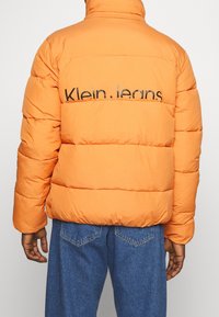 Pomarańczowa puchowa kurtka z pikowanym wzorem i czarnym logo "Klein.jeans" na plecach. Model ma na sobie niebieskie jeansy.