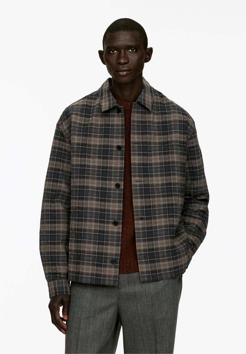 Homme portant une veste à carreaux bleue et marron sur un pull couleur rouille avec un pantalon gris, se tenant contre un fond blanc uni.
