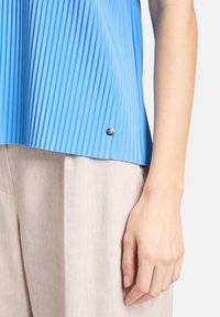 Niebieska plisowana bluzka o pionowej fakturze, z metalicznym akcentem na dole. Połączona z jasnobeżowymi spodniami z tkaniny.