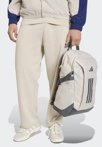 Pantalones deportivos de color beige claro, zapatillas a juego beige con tres rayas, y una mochila de color claro con tres rayas oscuras y detalles grises.