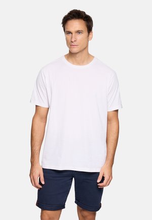 Hombre con camiseta blanca lisa de manga corta y pantalones cortos azul marino remangados, de pie frente a un fondo claro y sencillo.