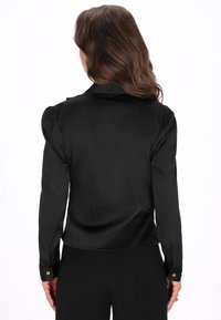 Zwarte satijnen blouse met lange mouwen, schouderplooien en gouden knopenaccenten op de manchet. Gladde textuur, getailleerde pasvorm en subtiele glans.