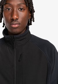 Black jacket with a high collar, featuring a mix of smooth and fleece textures, zippered front, and contrasting fabric accents on shoulders.
