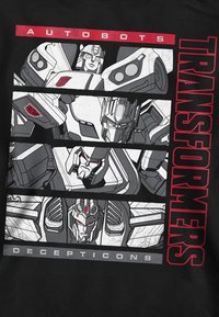 Schwarzes T-Shirt mit Comic-artigen Illustrationen von robotischen Charakteren, beschriftet mit "AUTOBOTS" und "DECEPTICONS" in fettem roten Text.