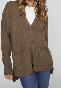 Ruskeasta neulepuvusta, jossa on V-kaula-aukko, neljä nappia ja kaksi etutaskua. Ribattua tekstuuria ja pidempi takapäällinen. Päällä valkoinen toppi.