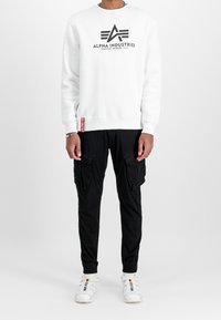 Sweat-shirt blanc avec logo "Alpha Industries" en noir, associé à un pantalon cargo noir avec multiples poches et des baskets blanches.