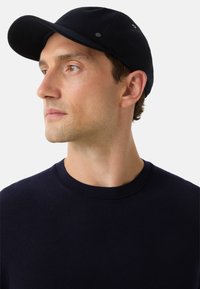 Námořní čepice se strukturovaným designem, s křivým štítkem a dvěma kovovými akcenty po stranách, nošená s tmavým svetr.