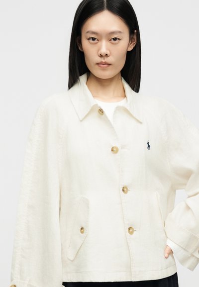 Mladá žena s rovnými čiernymi vlasmi nosí prehnane veľkú bielu bundu na gombíky s klopovými vreckami a malým vyšívaným logom na hrudi.