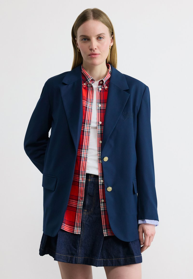 Γυναίκα που φοράει μπλε σακάκι πάνω από κόκκινο καρό πουκάμισο και λευκό τοπ, συνδυασμένα με φούστα από τζιν, στέκεται μπροστά σε απλό ανοιχτόχρωμο φόντο.