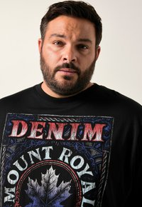T-shirt noir avec un grand design graphique mettant en avant "DENIM" en rouge et "MOUNT ROYAL" en blanc, entouré d'un motif de feuilles bleu et blanc.