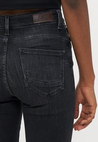 Tumšas denim bikses ar pieguļošu dizainu, kas aprīkotas ar brūnu ādas etiķeti aizmugurējā jostā un noapaļotu, izšūtu aizmugurējo kabatas detaļu.