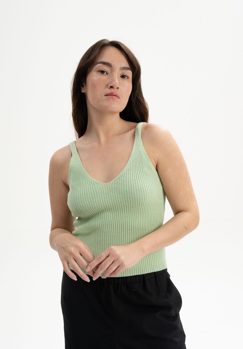 Helles grünes, geripptes Tanktop mit V-Ausschnitt, schmalen Trägern und einer figurbetonten Silhouette. Getragen mit schwarzen Hosen, die ein strukturiertes Strickmuster zeigen.