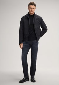 Giacca isolante nera con colletto alto, indossata sopra un maglione nero. Jeans in denim scuro e scarpe in pelle nere completano l'outfit.