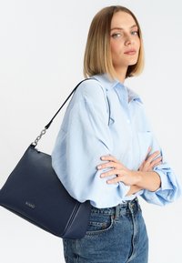 Borsa a tracolla in pelle navy con superficie testurizzata, caratterizzata da un design elegante e una tracolla accentuata da catena, indossata sopra una camicia azzurra chiara e jeans in denim.