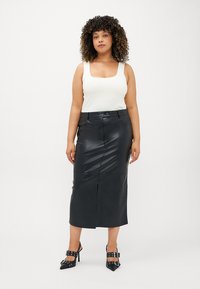 Μαύρη φούστα από ψεύτικο δέρμα με μήκος μέχρι το γόνατο, μπροστινή σχισμή και ομαλή υφή. Συνδυασμένη με εφαρμοστό λευκό τι-σερτ και ψηλοτάκουνα παπούτσια.