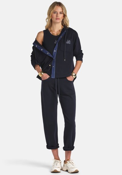 Szőke nő sötétkék kapucnis pulóverben, hozzáillő nadrágban, fehér sportcipőben, arany karkötőkkel és nyaklánccal, zsebeit markolva áll.