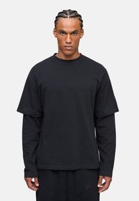 Schwarzes, übergroßes T-Shirt, das über einem Langarmoberteil getragen wird, aus Baumwolle gefertigt, mit Rundhalsausschnitt und subtilen Nähdetails.