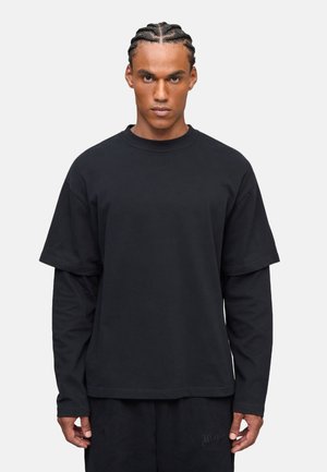 Schwarzes, übergroßes T-Shirt, das über einem Langarmoberteil getragen wird, aus Baumwolle gefertigt, mit Rundhalsausschnitt und subtilen Nähdetails.