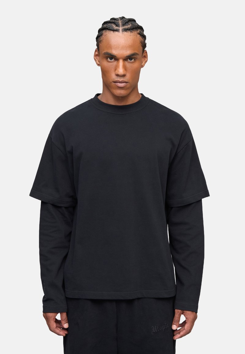 Schwarzes, übergroßes T-Shirt, das über einem Langarmoberteil getragen wird, aus Baumwolle gefertigt, mit Rundhalsausschnitt und subtilen Nähdetails.