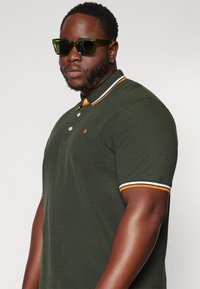 Zelené polo tričko s oranžovými akcenty na límci a okrajích rukávů, se třemi knoflíky. Model nosí oversized zelené sluneční brýle.