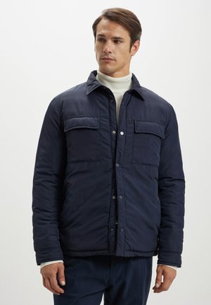 Giacca trapuntata blu navy con due tasche sul petto, chiusura frontale con bottoni a pressione e colletto. Presenta una texture liscia e una vestibilità rilassata.