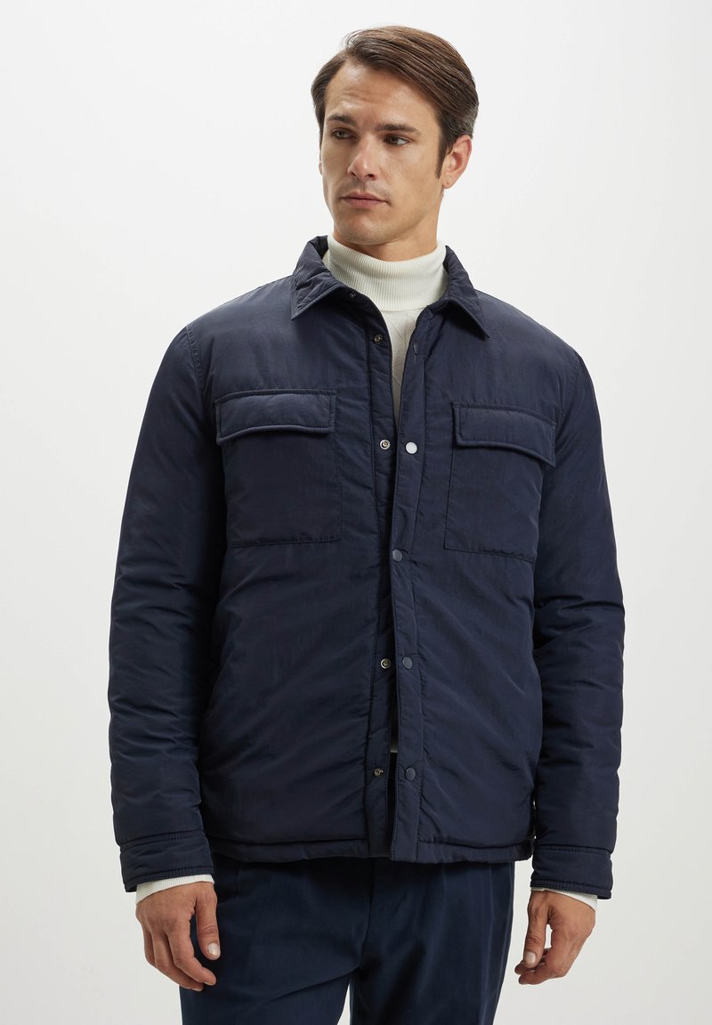 Giacca trapuntata blu navy con due tasche sul petto, chiusura frontale con bottoni a pressione e colletto. Presenta una texture liscia e una vestibilità rilassata.
