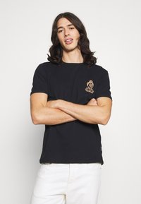 T-shirt de algodão preto com mangas curtas, apresentando um emblema dourado bordado no lado esquerdo do peito. Tecido com textura fina, corte regular.