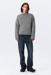 Jeune homme avec des cheveux bruns de longueur moyenne, portant un pull en maille gris, un jean bleu foncé avec un porte-clés et des chaussures noires, debout.
