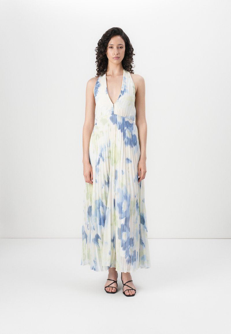 Lange, geplooide jurk met een V-hals, voorzien van aquarel bloemenpatronen in blauw en groen op een lichte achtergrond. Zwarte, strapsandalen.