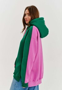 Preškrtnutá oversized mikina v zelenej a ružovej farbe. Obsahuje veľkú kapucňu, dlhé rukávy a rebrované manžety. Mäkký zmesový bavlnený materiál.