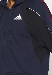 Primer plano de una sudadera con cremallera de Adidas en azul marino y negro, con el logotipo blanco en el pecho y una franja roja en la manga.