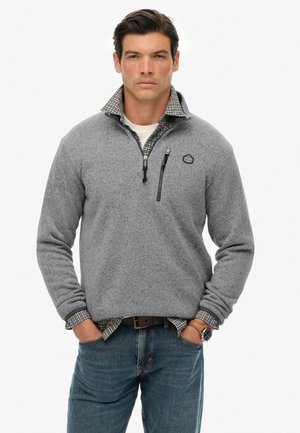 Grå fleecepullover med halv lynlås, der har en ternet krave, brystlomme og en tekstureret overflade. Bæres over en hvid skjorte og jeans.