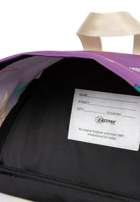 Interiér fialové tašky s černým podšívkou a štítkem s prostorem pro osobní údaje. Zajímavý design zahrnuje barevné grafické vzory.