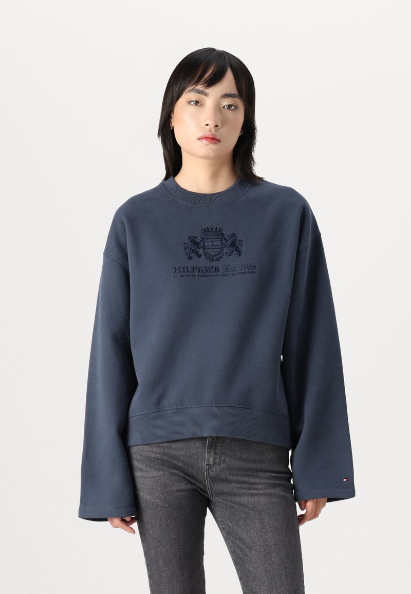 Sweatshirt bleu marine avec un col rond, des manches larges et un logo brodé représentant un blason à l'avant. Texture lisse.
