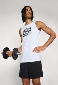 Camiseta sin mangas blanca con texto gráfico negro "JACK & JONES PERFORMANCE", combinada con pantalones cortos negros y sosteniendo una mancuerna.