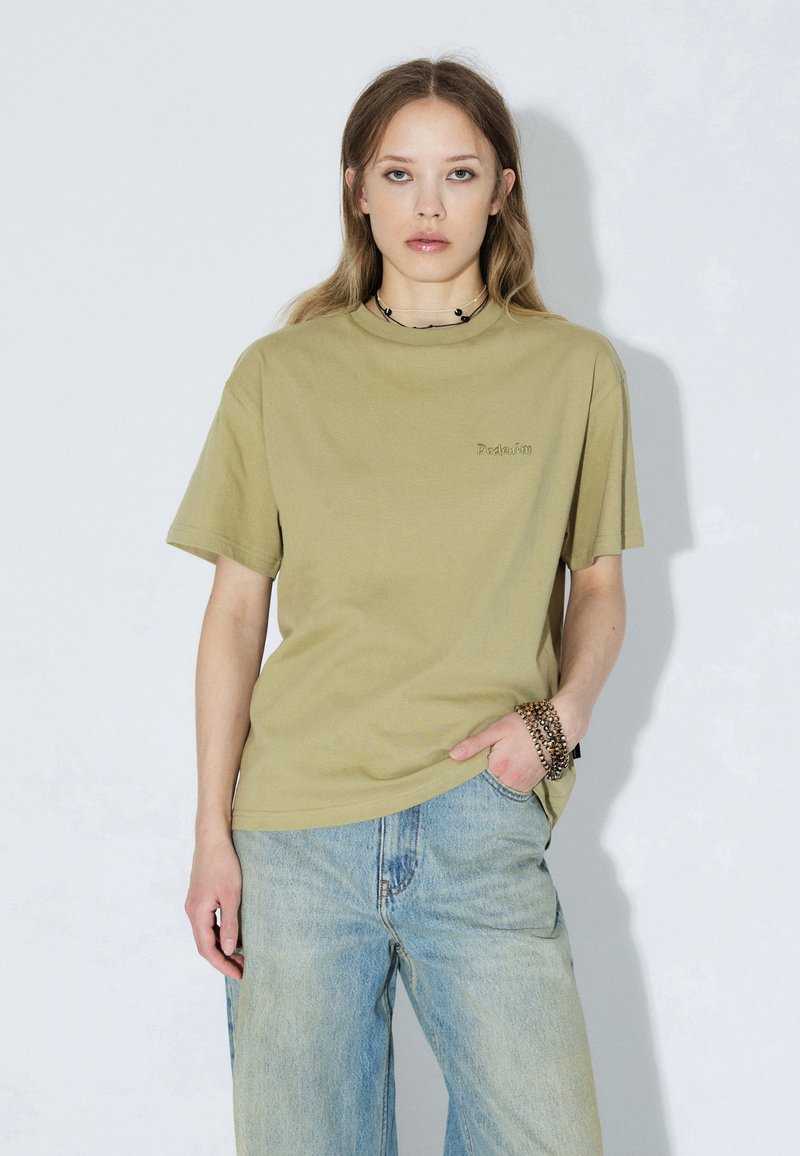 T-shirt di cotone verde oliva con scollo rotondo, maniche corte e ricamo discreto "De:Ad:Um" sul petto, abbinata a jeans denim azzurri.