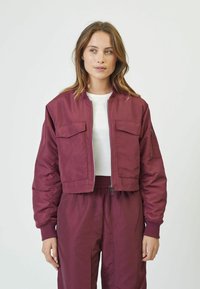 Burgundy cropped bomber jacket with zipped front, two chest pockets, and ribbed cuffs, worn over a white shirt and matching trousers.
