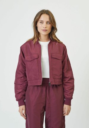 Burgundy cropped bomber jacket with zipped front, two chest pockets, and ribbed cuffs, worn over a white shirt and matching trousers.