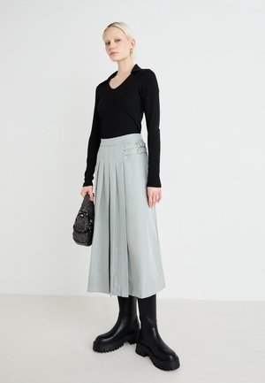 Femme blonde portant un haut noir à manches longues, une jupe plissée gris clair avec des boucles, des bottes noires épaisses, tenant un petit sac à main noir.