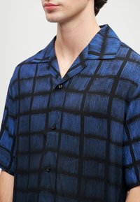 Mann som har på seg en kortermet knappskjorte med blå og svart rutemønster, med camp krage, avkortet ved nakken og øvre torso.