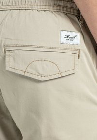 Pantalones cortos de algodón beige que cuentan con un bolsillo trasero con detalles de costura y una etiqueta blanca que dice "Reell est. 1997."