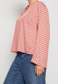 Red and white patterned long-sleeve shirt featuring a scallop design, V-neckline, and relaxed fit, worn with blue jeans.